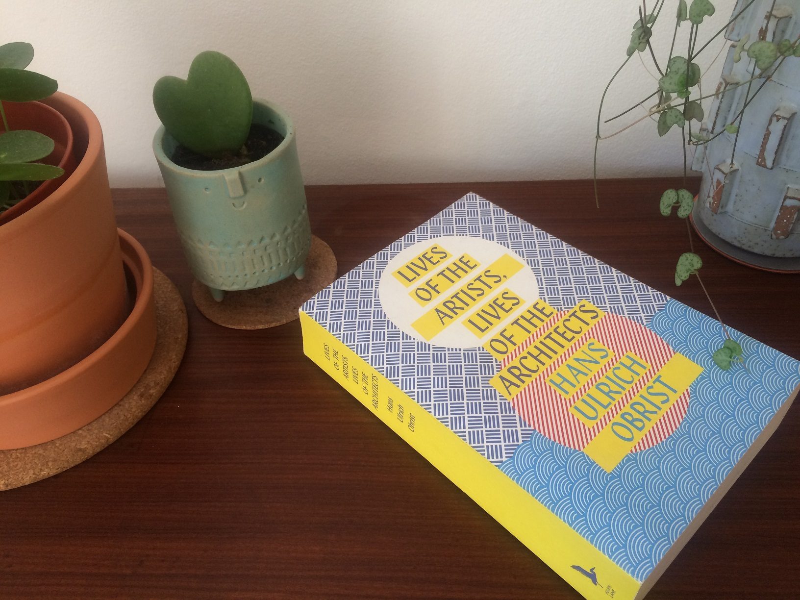 Photograph of the book Lives of the Artists, Lives of the Architects by Hans Ulrich Obrist on a sideboard surrounded by plant pots.