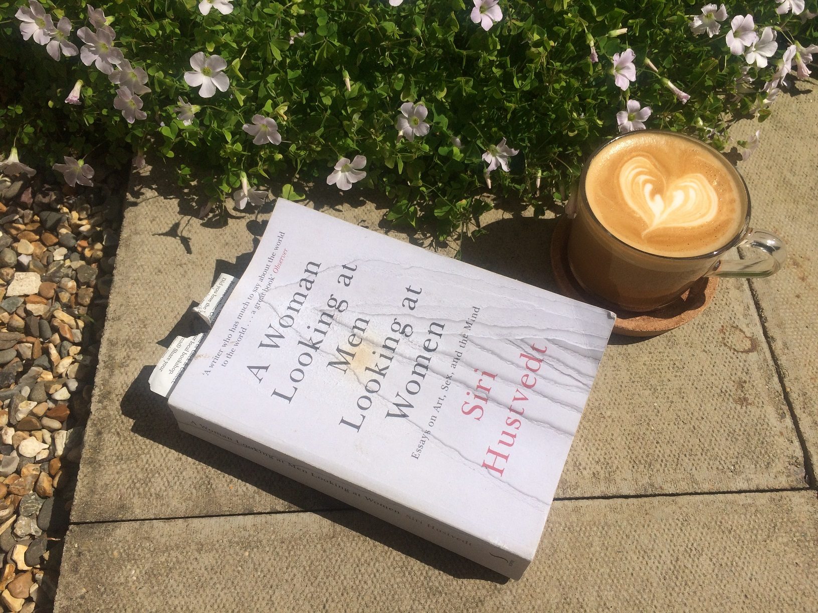 A photograph of the book 'Women LOoking at Men' by author Siri Hustvedt on a garden paving stone with a coffee and some plants.