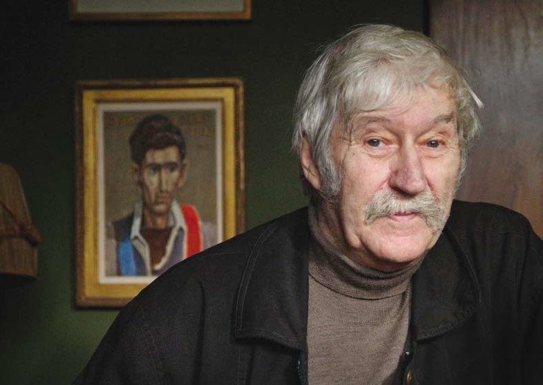 Photograph of a man with white hair and moustache in front of a portrait of a young man with dark hair.
