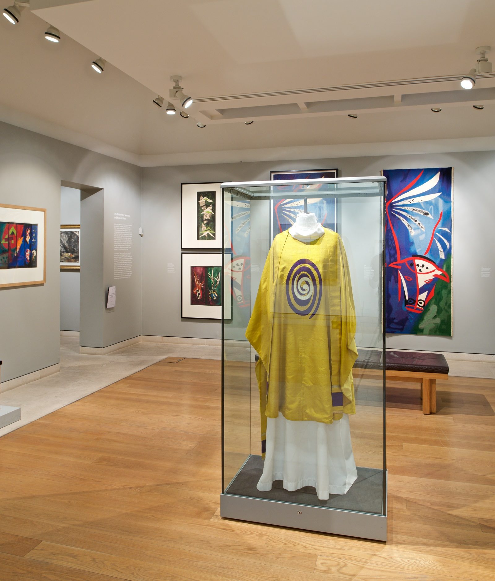 View of gallery showing abstract works of art on the wall. In the centre is a glass case containing a mannequin wearing a yellow vestement with abstract oval design on its chest