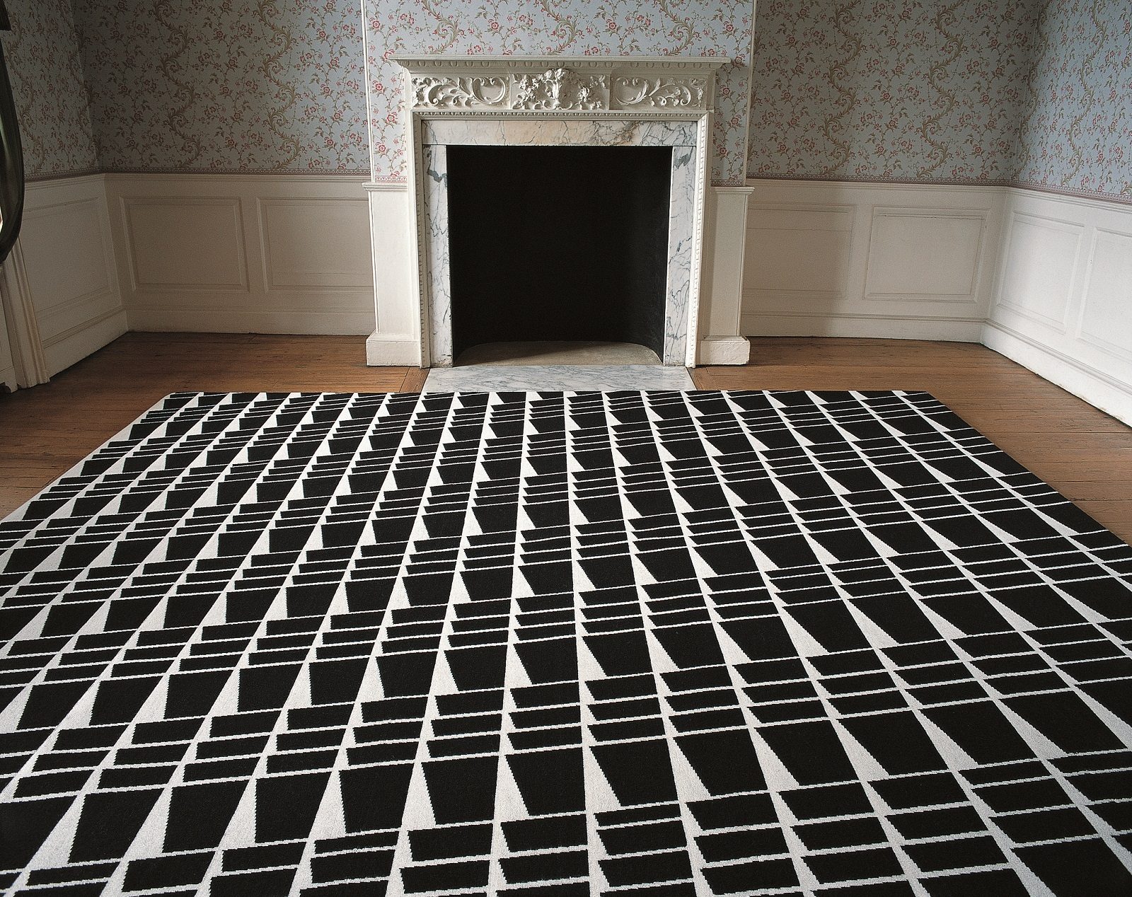 A black and white rug in a geometric pattern laid out in front of a period fireplace in a room with floral wallpaper