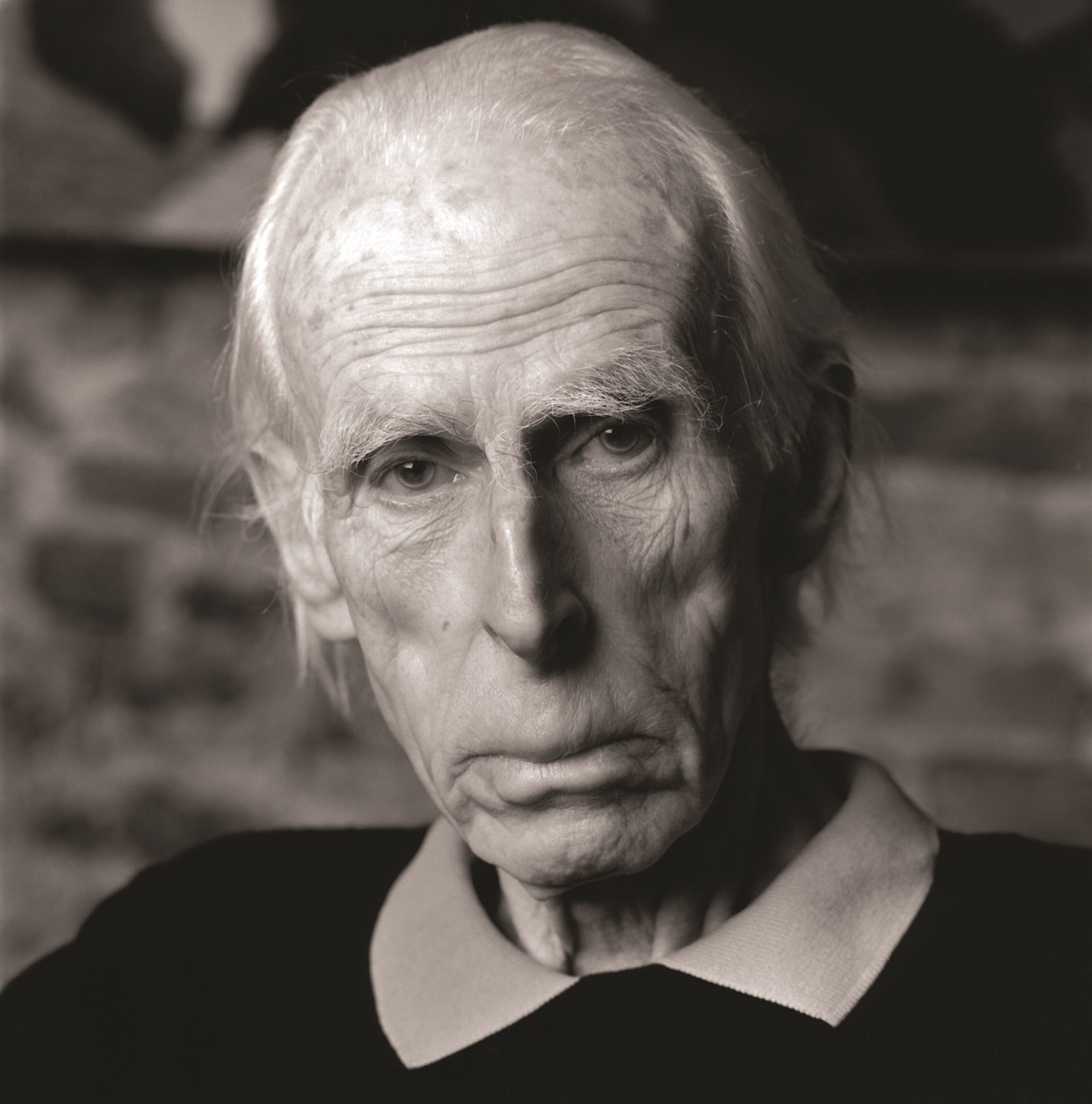 Black and white photograph showing the head and shoulders of a man with thinning white hair staring directly at the camera