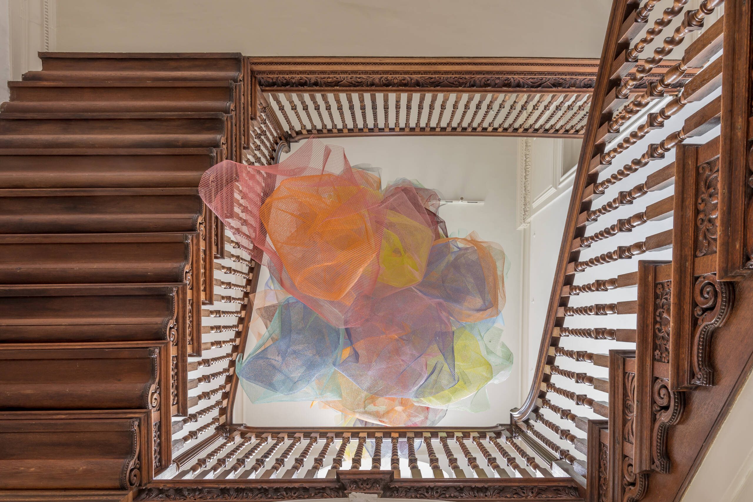 Photograph of a coloured mesh installation in a stairwell shot from below with the wooden staircase and balustrade all around