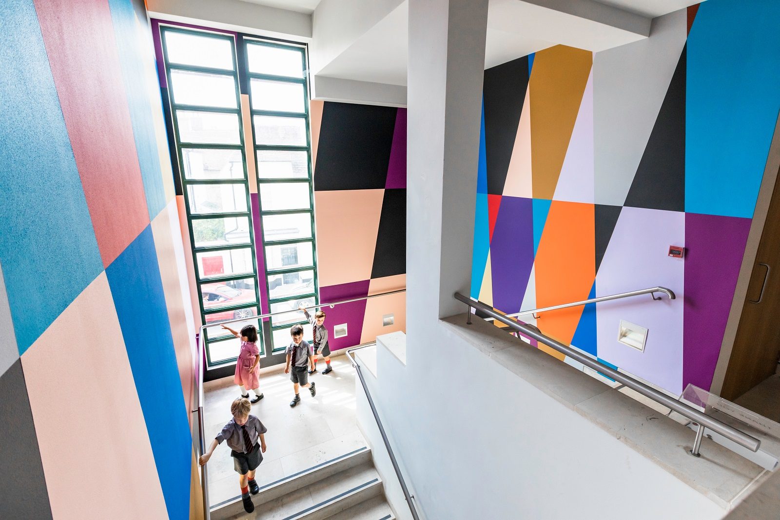 Three children in school uniform descend a staircase decorated with a brightly coloured geometric mural